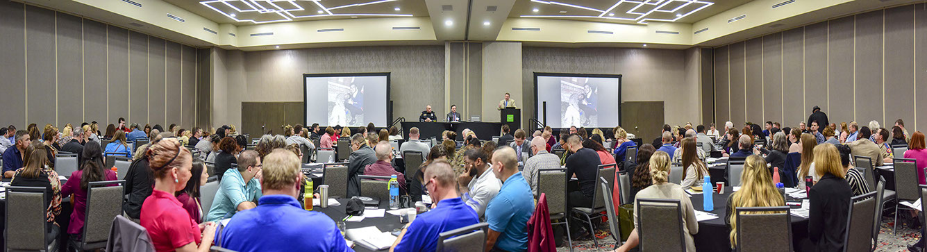 Photo of a conference room where several people sit at tables, two people sit in the front of the room, and one presents to the audience.