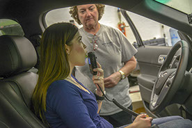 Photo of a young woman in a car blowing into the ignition interlock device while another woman standing outside the car watches.