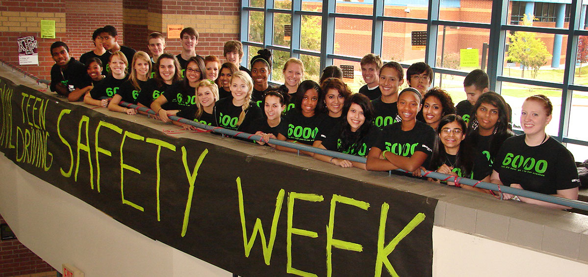 Photo of a group of teenagers, wearing  shirts that say 6000, standing against a railing. A sign says: Teen Driving Safety Week.